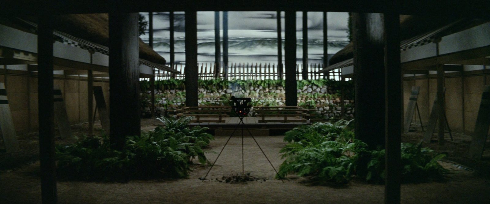 An abandoned wooden house open to the sky: rotting beams frame an overgrown courtyard, a painted grey sky beyond
