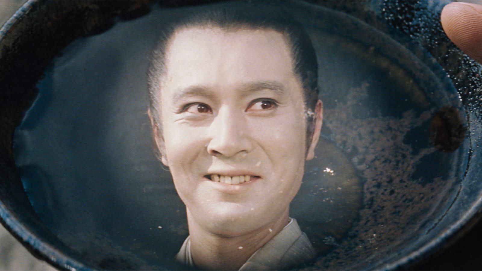 A smiling young man’s face reflected in the tea at the bottom of a dark bowl held in a hand