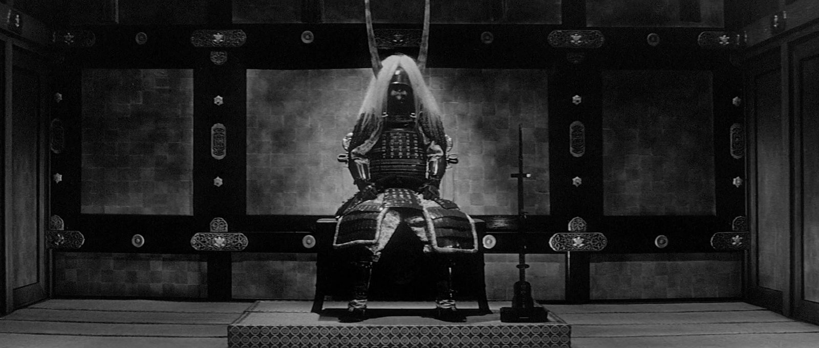 An empty suit of samurai armor sits on a dais at the end of a great hall, in black and white