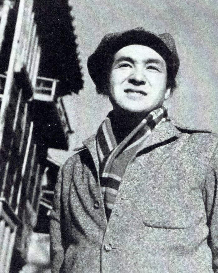 Masaki Kobayashi in a beret and a striped scarf, smiling, photographed from below in front of a wooden building