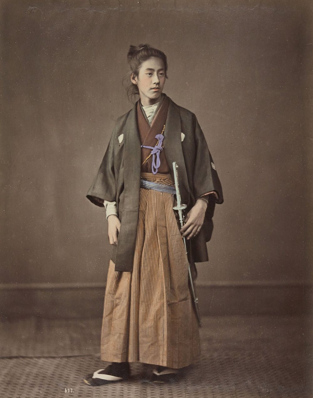 A young man in a striped hakama and a dark coat, his hair drawn up into a topknot, holding a sword at his side, in a hand-colored studio portrait