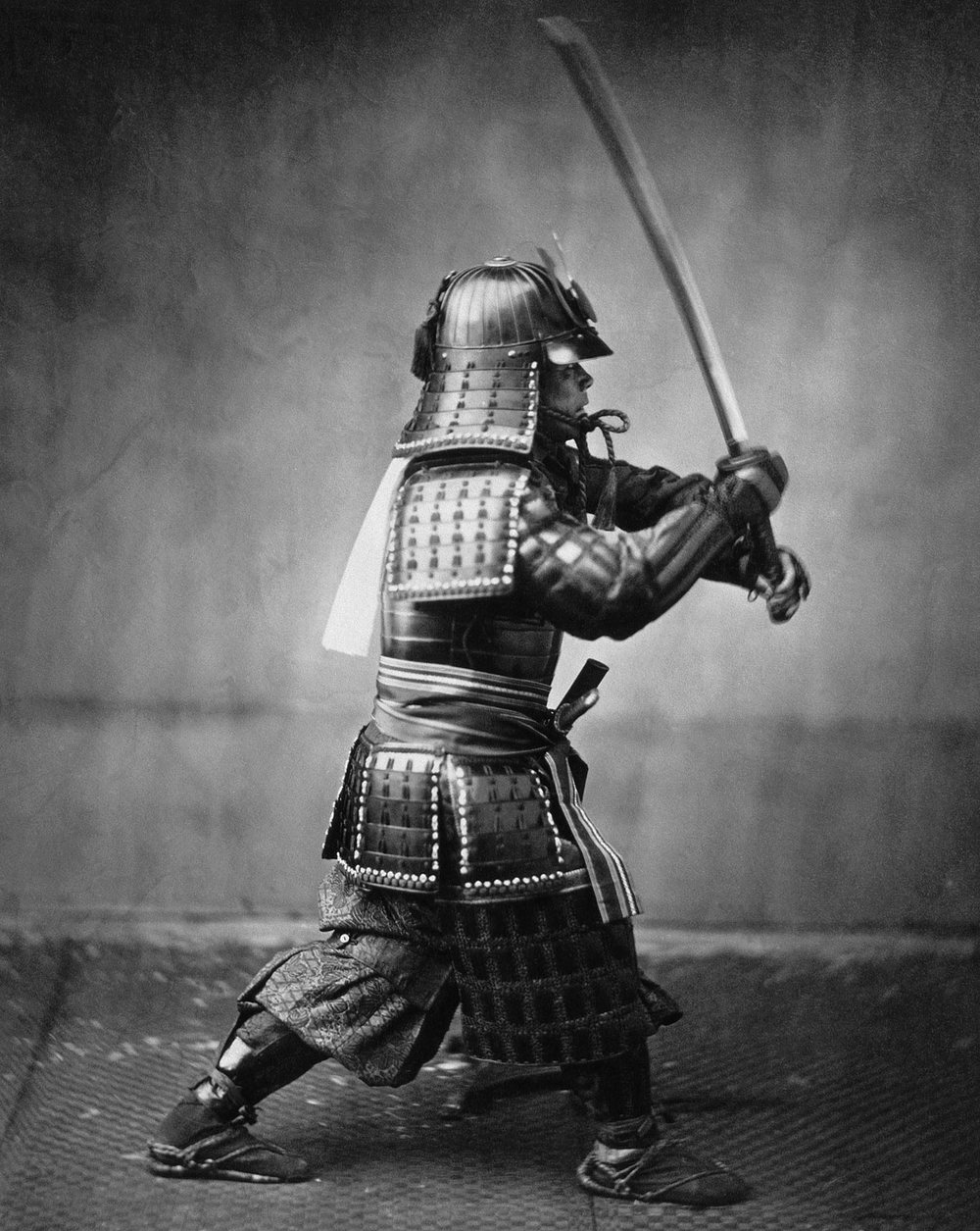 A samurai in full armor and a lacquered helmet crouches with a long sword raised above his head, against a plain studio backdrop