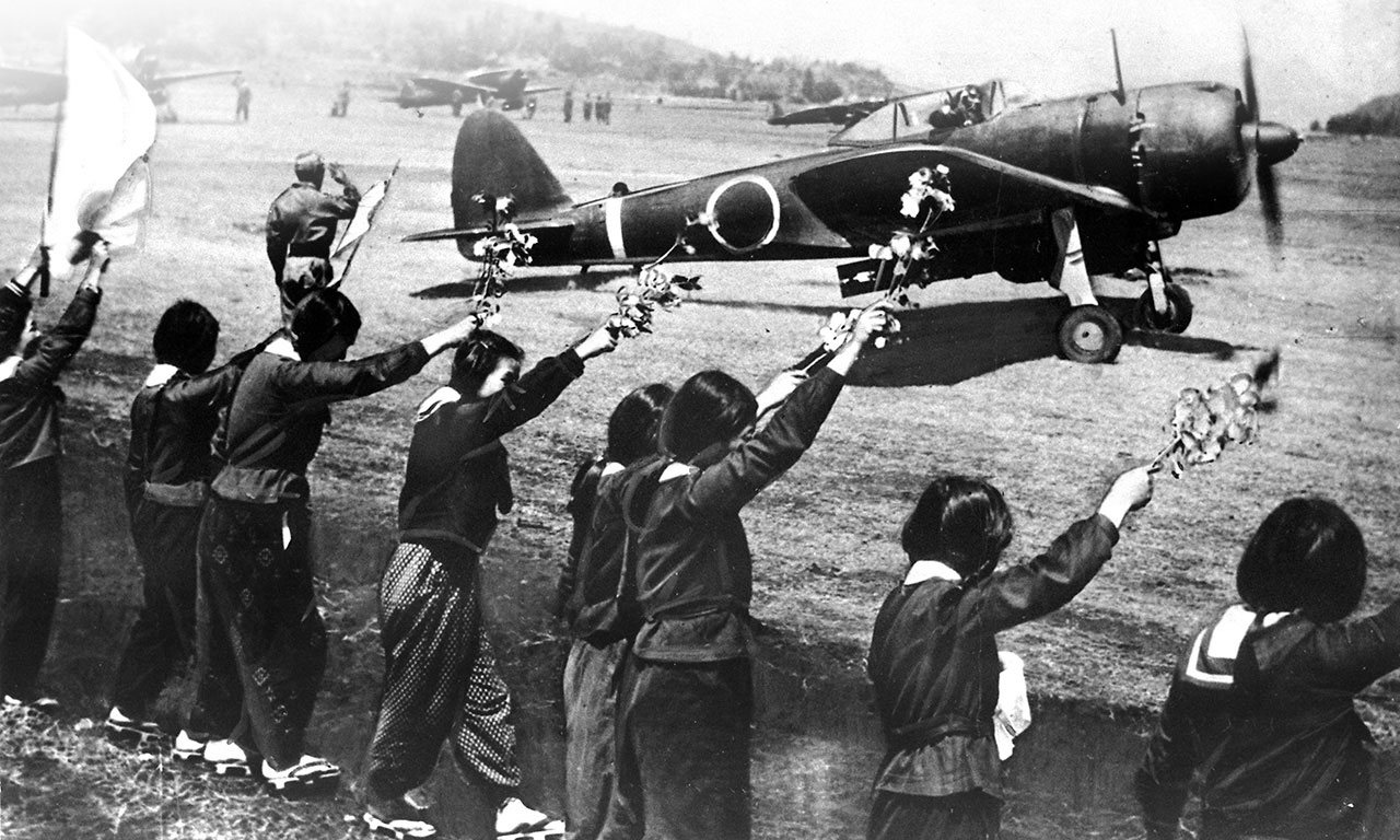 Schoolgirls in dark uniforms wave branches of cherry blossom at a fighter plane taking off past them