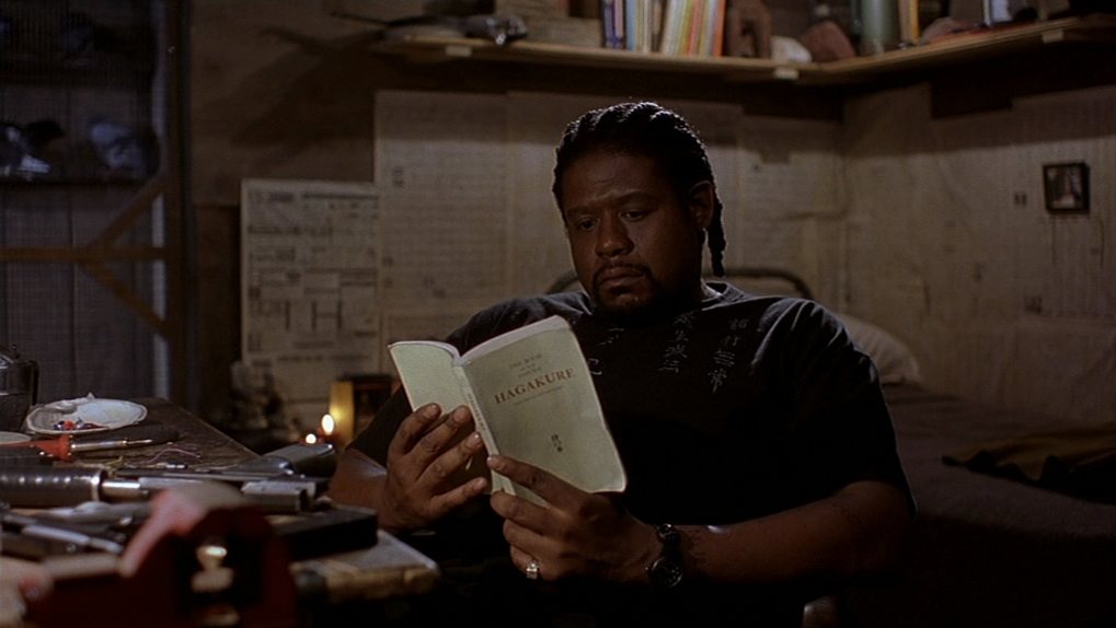 Ghost Dog, a heavyset man with braided hair, sits at a cluttered workbench in his rooftop shack reading a paperback whose cover says Hagakure