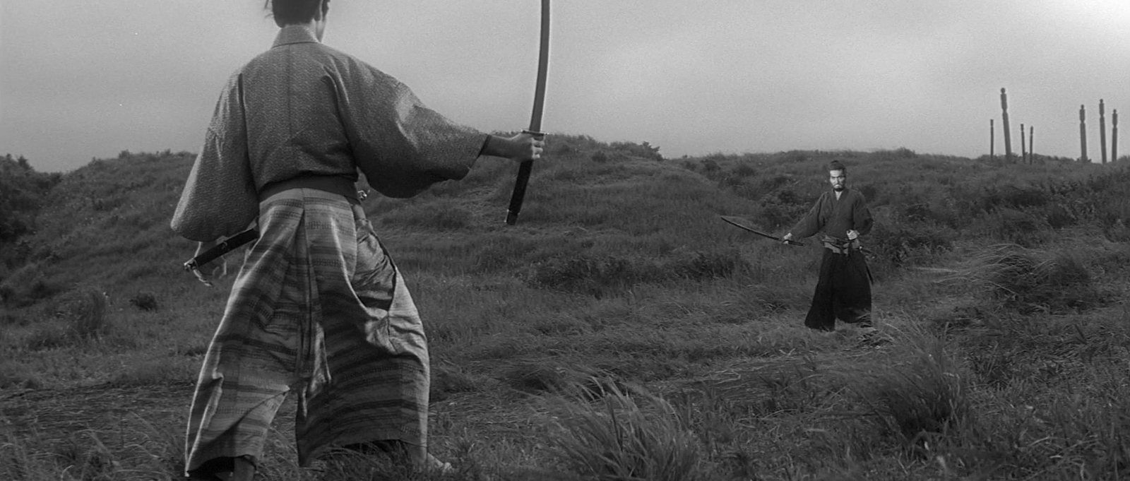 On a windswept hill of long grass, a samurai seen from behind, his topknot tied high and his sword raised, faces another swordsman across the field, with tall grave markers on the ridge beyond