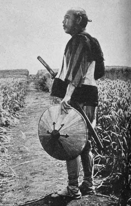Takashi Shimura as Kanbei, his head shaved like a monk’s, standing on a path through tall grass with a straw hat in one hand and his swords at his side