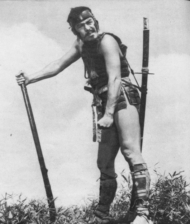 Toshirō Mifune as Kikuchiyo, bare-legged in a field of grass, leaning on a sword longer than he is tall