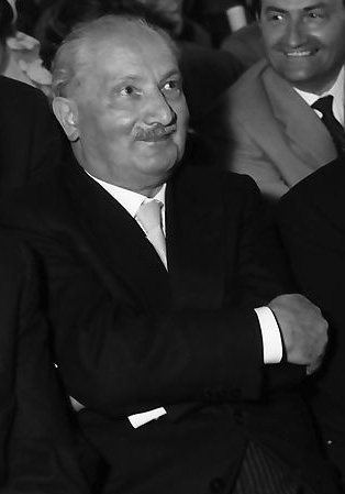 Martin Heidegger in 1960, seated in a dark suit at a public ceremony, smiling slightly