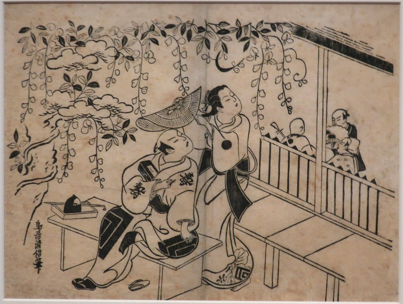 A black-and-white woodblock print: under hanging wisteria a young man sits on a bench beside a teahouse while a young woman standing behind him lifts a wide straw hat over his head; onlookers watch from the veranda