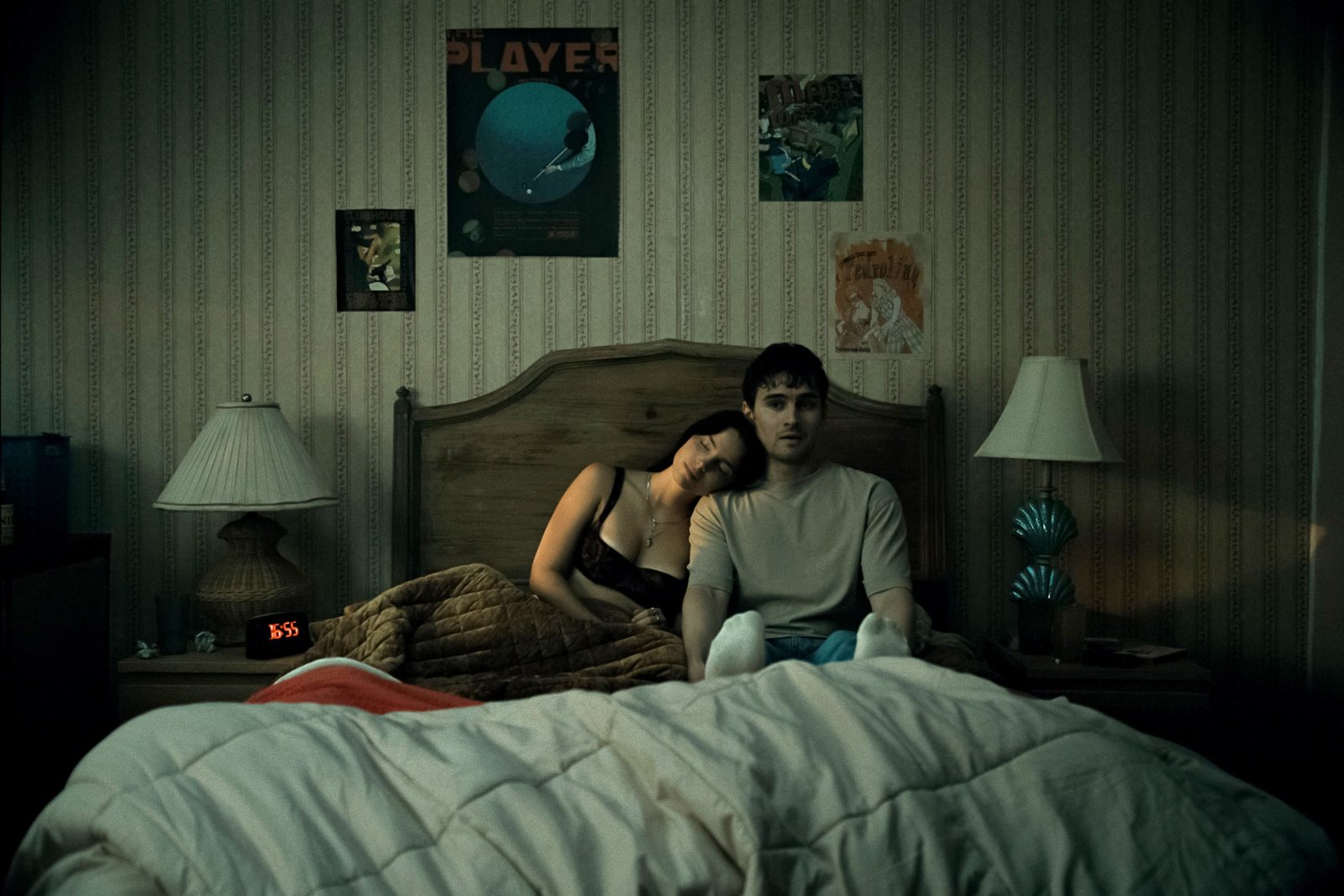Nikki and Bear sitting up together in a narrow bed at night under a striped wallpaper, a lamp glowing on each side; she leans her head on his shoulder while he stares ahead