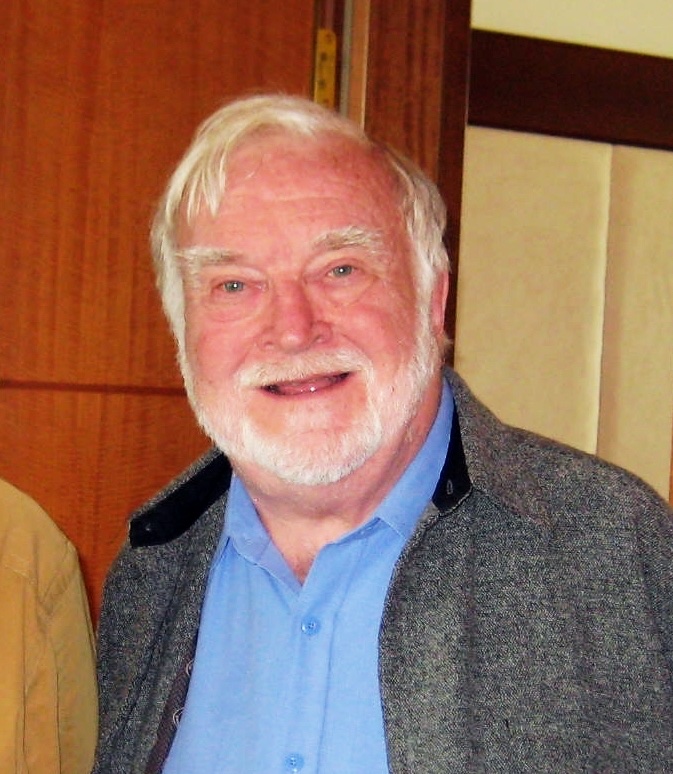 Mihaly Csikszentmihalyi, white-haired and bearded, smiling, in a blue shirt and grey jacket